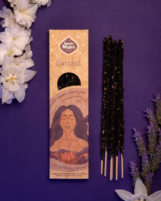 Sahumerio 5 Elementos Éter artesanal con varillas gruesas, presentado en envase decorativo, acompañado de flores sobre fondo púrpura.
